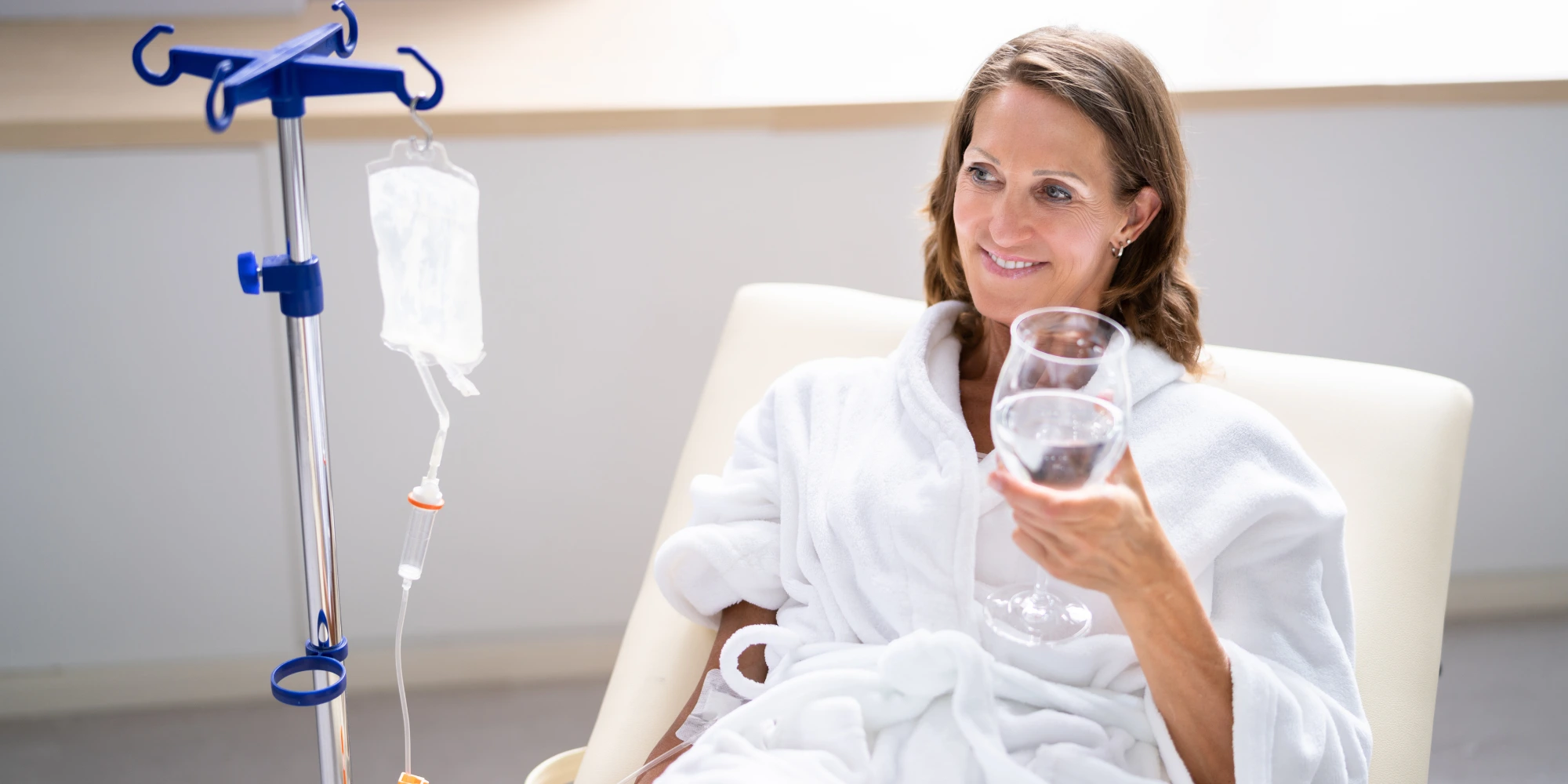 A person in a white robe receiving glutathione IV therapy, holding a glass of water, with a serene and radiant expression, symbolizing wellness and rejuvenation.
